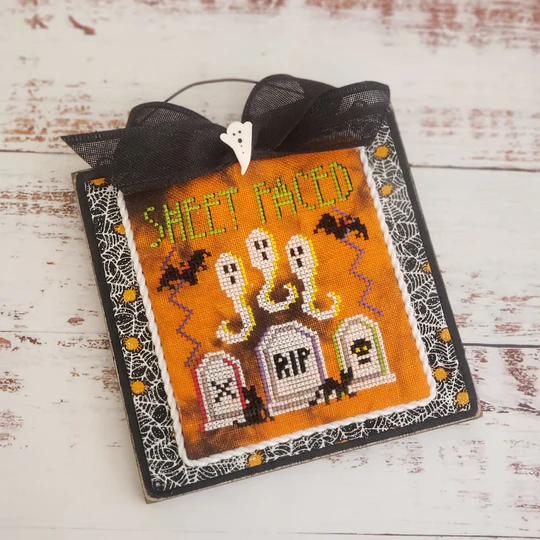 Halloween-themed cross-stitched piece with ghosts, tombstones, and text on an orange background.