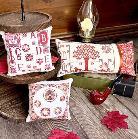 Three decorative throw pillows with red embroidered autumn-themed patterns on a white fabric, styled on a rustic wooden table with a candle, pipe, and leaves.