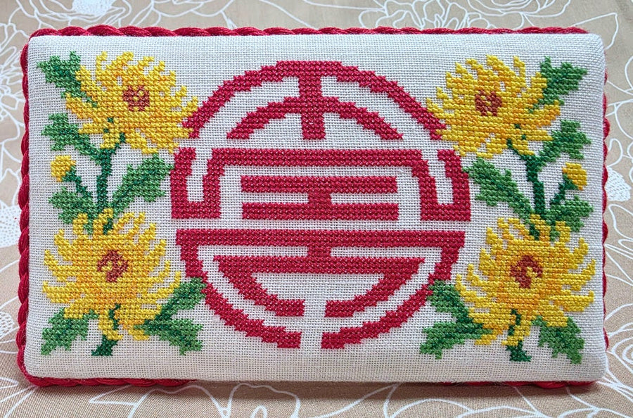 A rectangular embroidered cushion cover featuring a large red Chinese double happiness symbol in the center, flanked by yellow chrysanthemums with green leaves on a white embroidered fabric background with a red border.