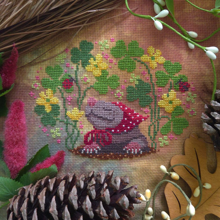 A close-up of a cross-stitch or needlepoint textile featuring a gray frog wearing a red polka-dot blanket, surrounded by green clover and yellow flowers. The fabric is styled with natural elements including pinecones, a pink flower, and a dried leaf.