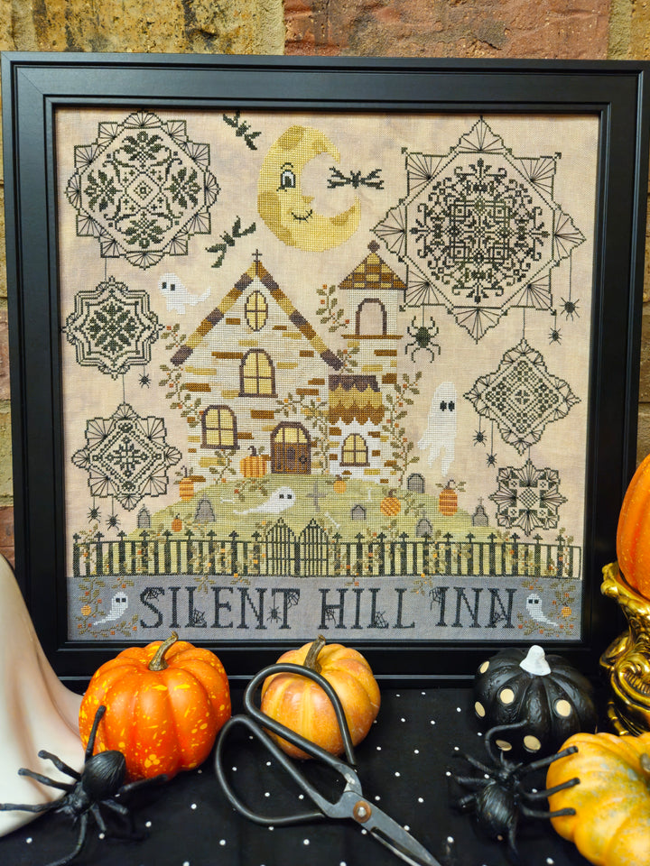 A framed cross-stitch artwork depicting a haunted house with the text 'SILENT HILL INN' at the bottom, displayed on a black surface with pumpkins, scissors, and spider decorations in a Halloween-themed setting.