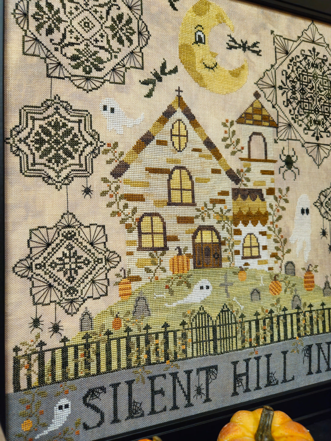 A framed cross-stitch sampler featuring a haunted house, ghosts, pumpkins, bats, a smiling crescent moon, and the words 'SILENT HILL IN' on a gray banner at the bottom.