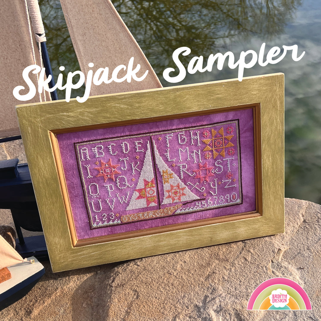 A framed needlework piece depicting a skipjack boat with quilt block motifs, displayed outdoors with water in the background.