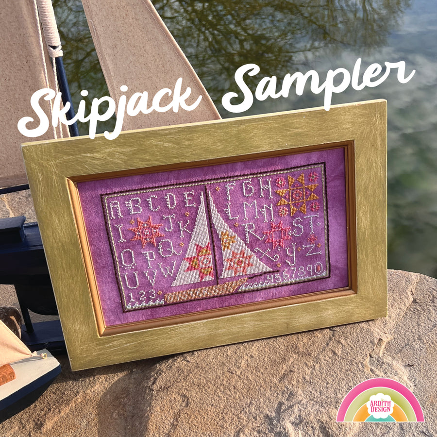 A framed needlework piece depicting a skipjack boat with quilt block motifs, displayed outdoors with water in the background.