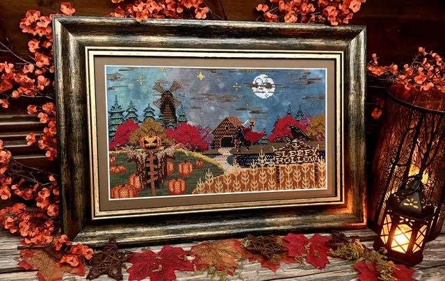 A framed autumn-themed needlepoint or cross-stitch artwork displayed on a rustic wooden surface, surrounded by red fall leaves, orange berries, and a lit lantern. The artwork depicts a moonlit countryside scene with a windmill, cabins, pumpkins, and the words 'Happy Fall Hollow.