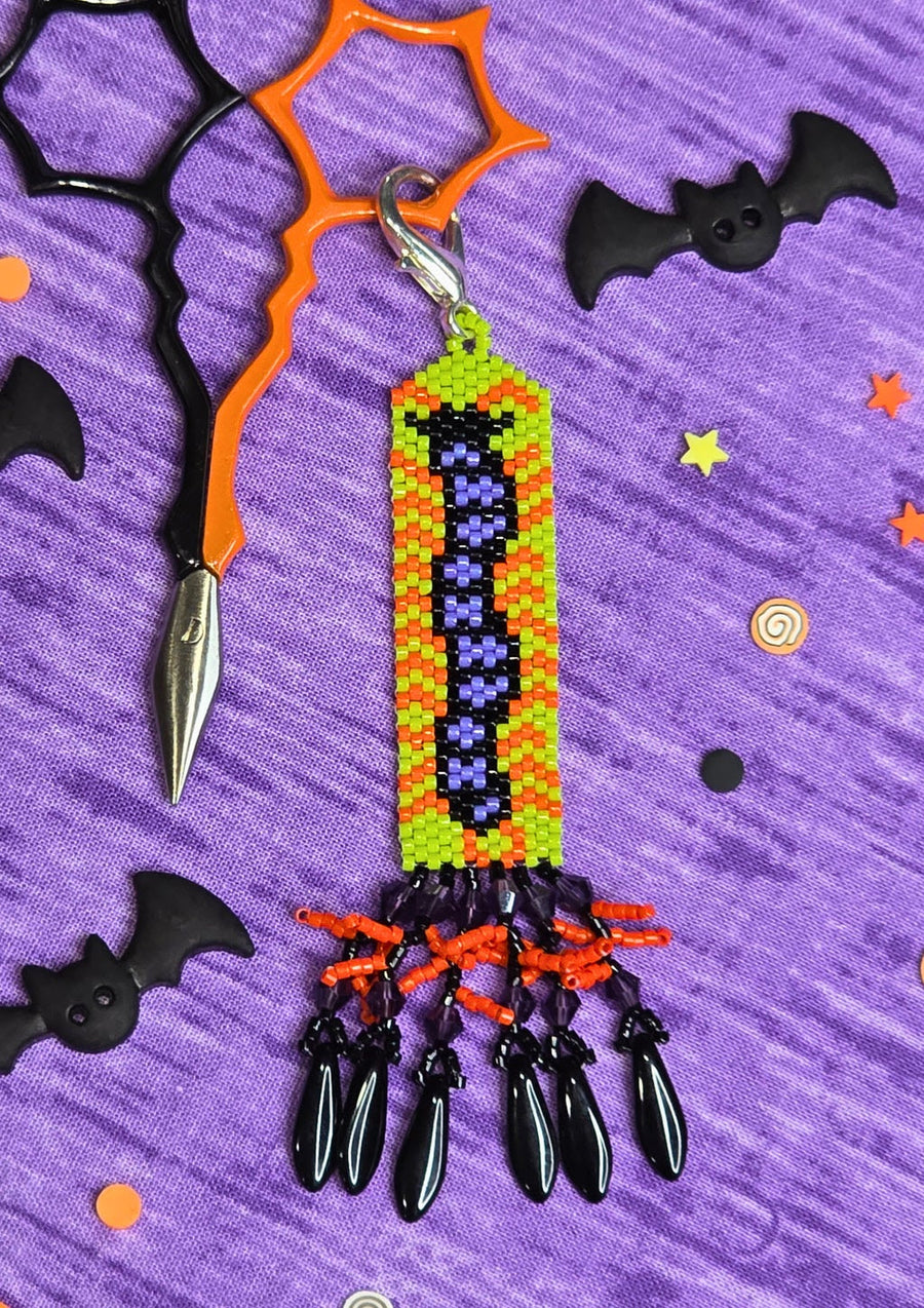 A Halloween-themed beaded keychain featuring a purple witch's hat on a green and yellow striped background, with dangling black teardrop charms. The keychain is styled on a purple textured background with black bat cutouts, orange scissors, and small star decorations.