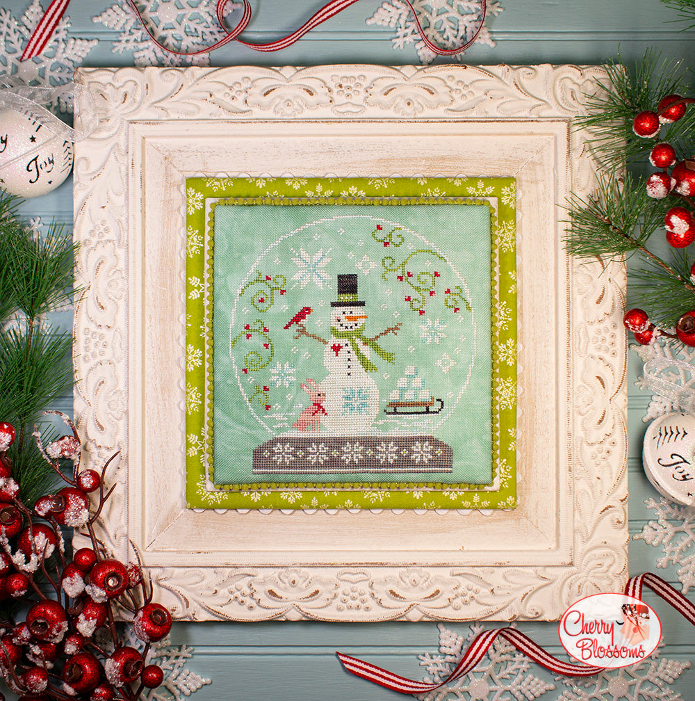 Framed cross-stitch artwork of a snowman in a snow globe with Christmas decorations around.