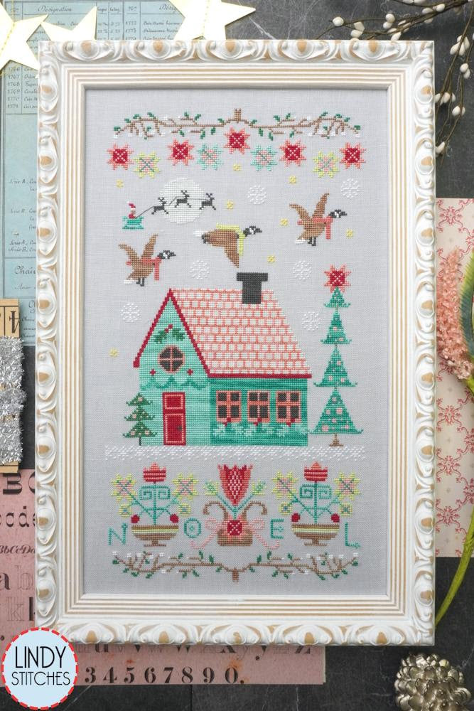 Framed cross-stitched design of a house with festive elements, featuring Lindy Stitches branding.