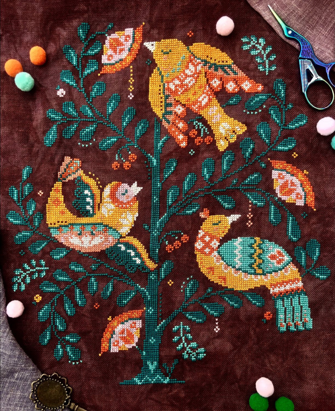 A finished cross-stitch or needlepoint artwork depicting three stylized birds perched on a tree, stitched on a dark brown fabric background. Scissors and colorful pom-poms are placed around the edges as styling props.