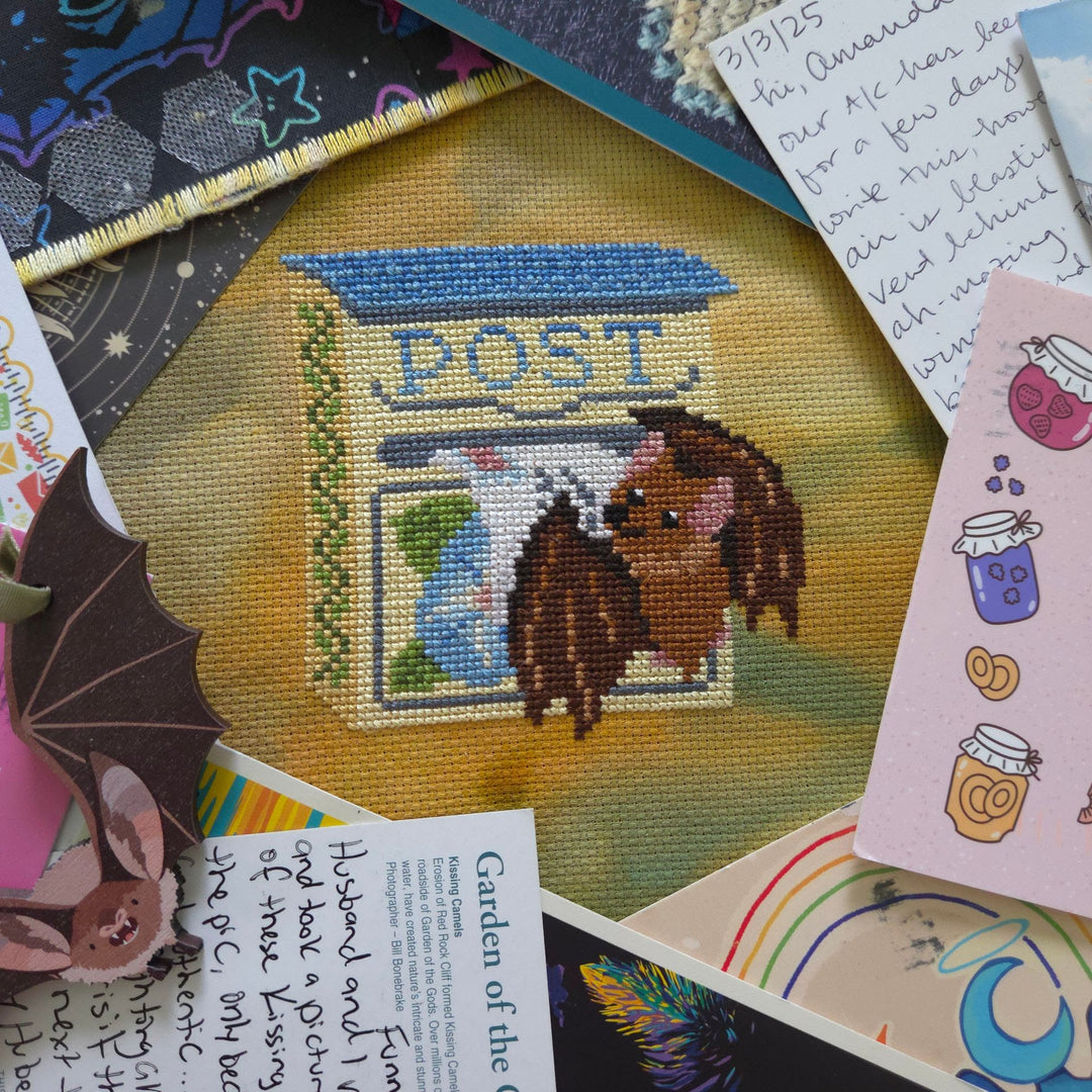 A close-up of a finished cross-stitch piece depicting a brown hedgehog standing in front of a blue and green post box with the word 'POST' stitched on top. The embroidery is displayed on a yellow fabric base and surrounded by decorative paper notes, a ruler, and other craft items.