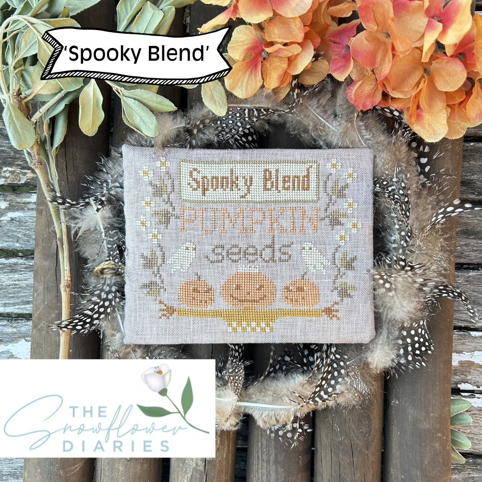 Cross-stitched panel with 'Spooky Blend Pumpkin Seeds' design, surrounded by autumnal elements on a wooden background.