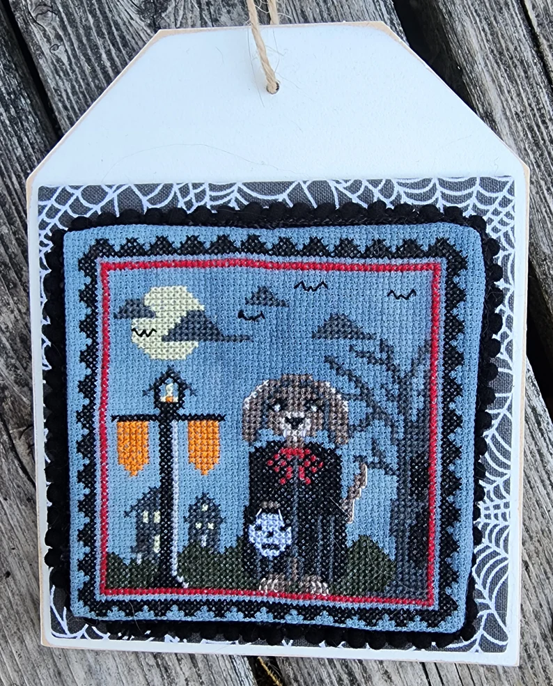 Decorative cross-stitched piece with a ghost and moon design on a wooden surface