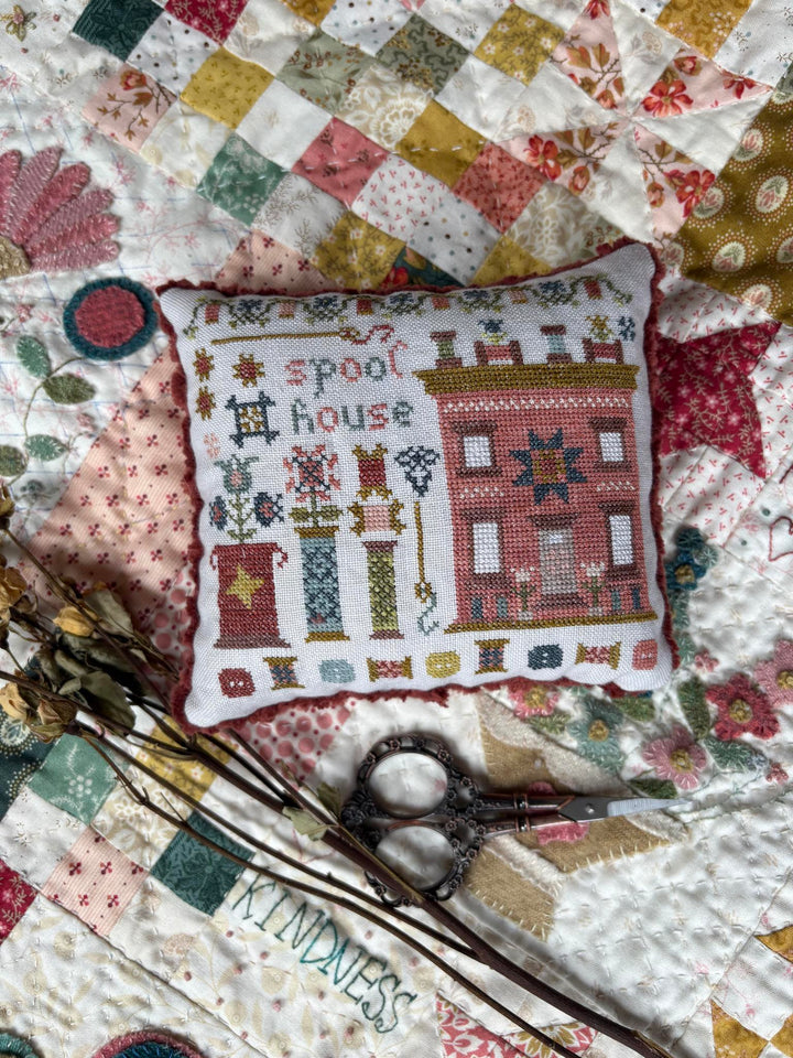 A square cross-stitch pillow with a 'Spool House' theme, featuring a red house in the shape of a spool and various spool motifs on a white fabric background, styled on a colorful patchwork quilt with scissors and dried flowers.