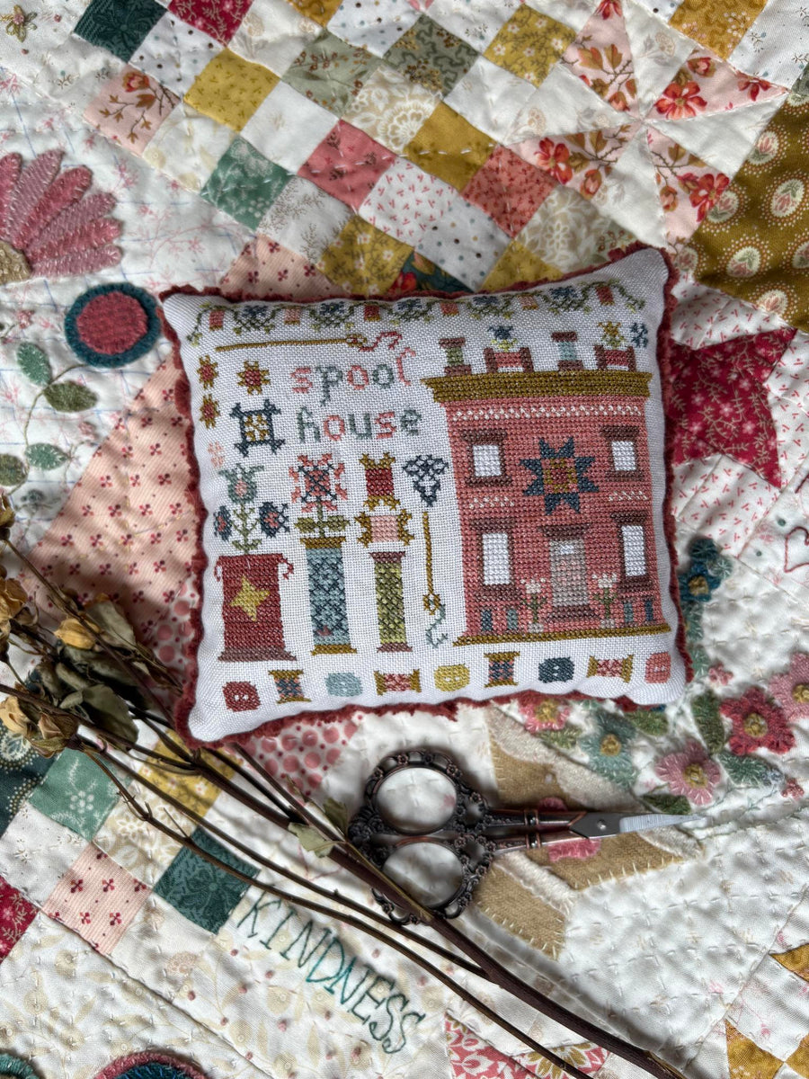 A square cross-stitch pillow with a 'Spool House' theme, featuring a red house in the shape of a spool and various spool motifs on a white fabric background, styled on a colorful patchwork quilt with scissors and dried flowers.