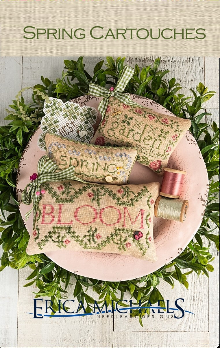Cross-stitched spring-themed cartouches on a pink plate with greenery and thread spools.