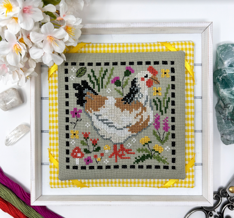 Framed cross-stitched artwork of a rooster with flowers and butterflies on a yellow checkered border.