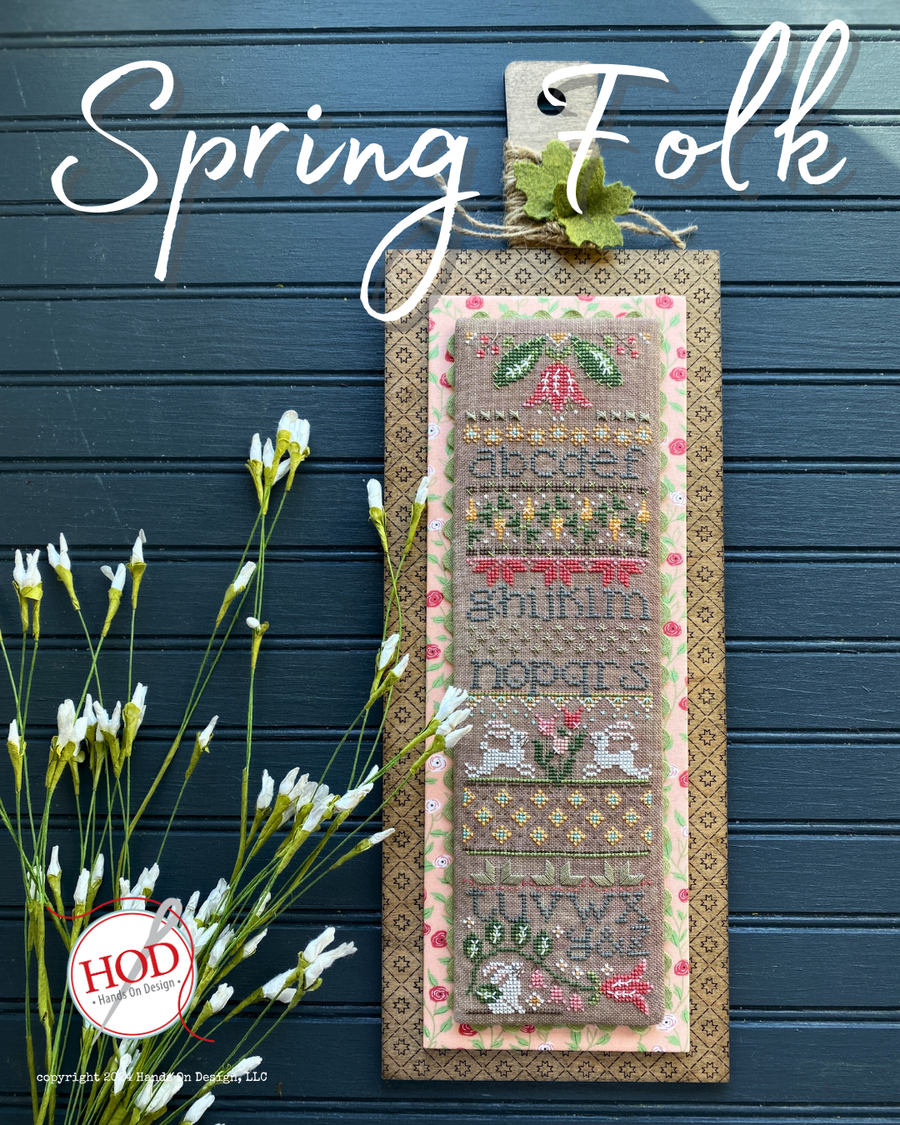 Cross-stitched panel with floral and leaf designs on a blue wooden background, featuring the brand 'HOD'.