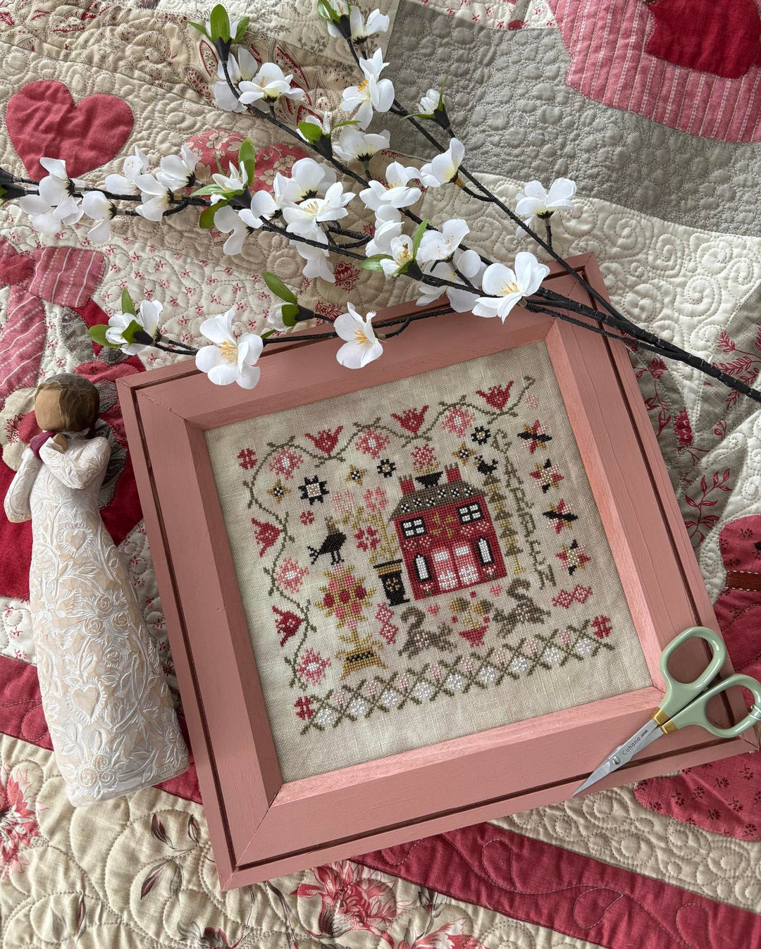 Framed cross-stitched piece on a quilt with floral branches and scissors.