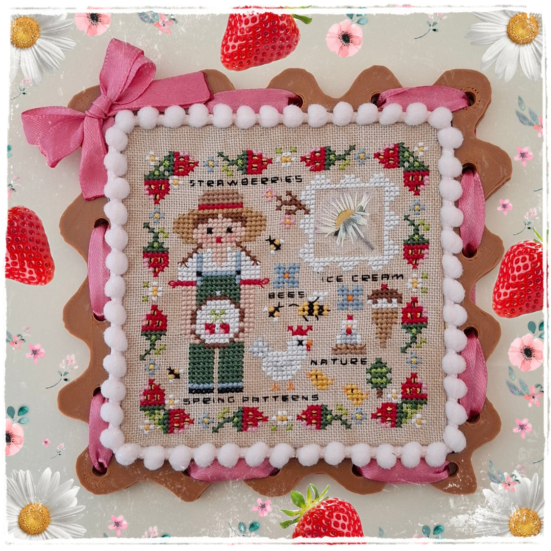 A square cross-stitch sampler featuring a strawberry-themed design with a girl, strawberries, flowers, and decorative text, framed by pink ribbon and white pom-pom trim on a floral background.