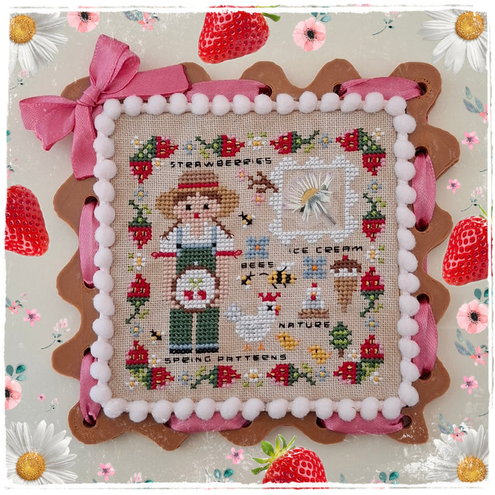 A square cross-stitch sampler featuring a strawberry-themed design with a girl, strawberries, flowers, and decorative text, framed by pink ribbon and white pom-pom trim on a floral background.