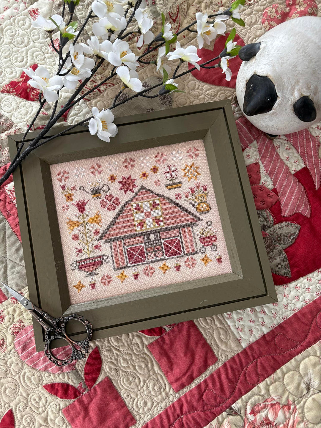 Framed cross-stitched design of a barn on a quilt with floral decorations.
