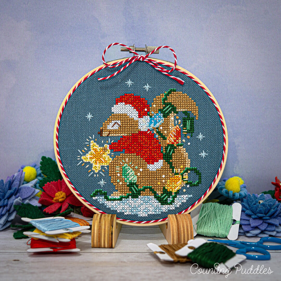 Cross-stitched squirrel in a Santa hat with a star and gift, surrounded by embroidery supplies on a wooden surface.
