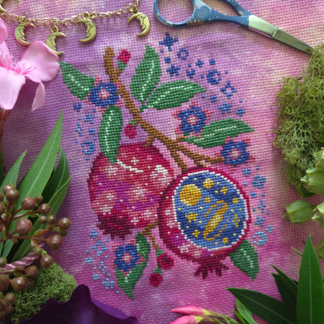 A close-up of a pink needlepoint canvas featuring a pomegranate design with green leaves, blue flowers, and a crescent moon and sun motif inside one pomegranate. The canvas is styled with jewelry, scissors, moss, and flowers around it.