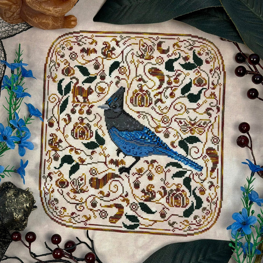 A square cross-stitch or needlepoint artwork featuring a blue bird at center, surrounded by autumn motifs including acorns, leaves, and small figures, laid flat on a light fabric background with decorative flowers and leaves around it.