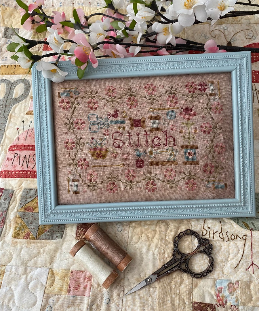 A cross-stitch pattern with the word 'Stitch' in the center, framed in a light blue frame, displayed on a quilt with a floral pattern in the background.