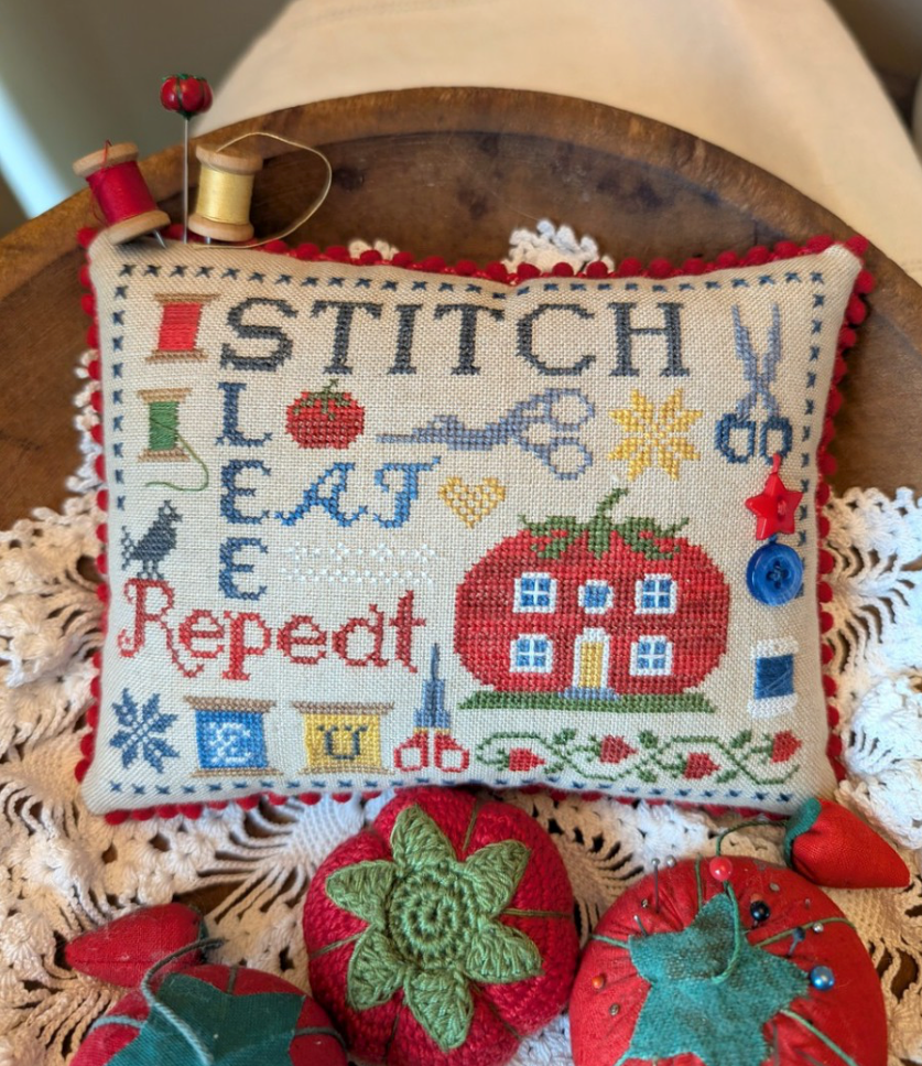 A rectangular cross-stitch pillow with a red pom-pom border, featuring the words 'STITCH LEAVE REPEAT' in embroidered letters alongside sewing-themed motifs like a house, scissors, and buttons. The pillow is styled on a wooden surface with spools of thread, a lace doily, and strawberry-shaped pin cushions.