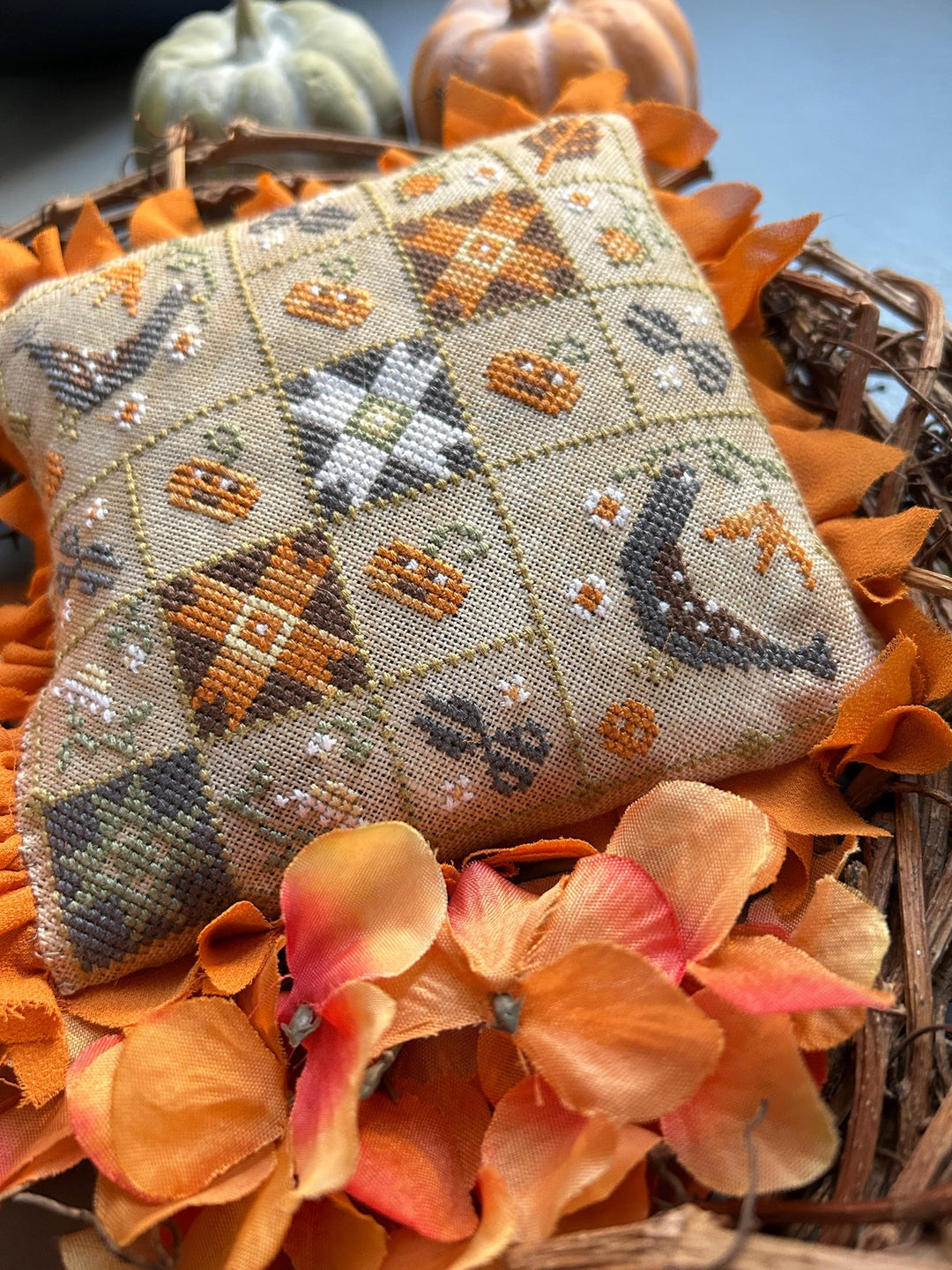 A square decorative pillow with a cross-stitch style autumn pattern, displayed on a wreath surrounded by orange leaves and small pumpkins.
