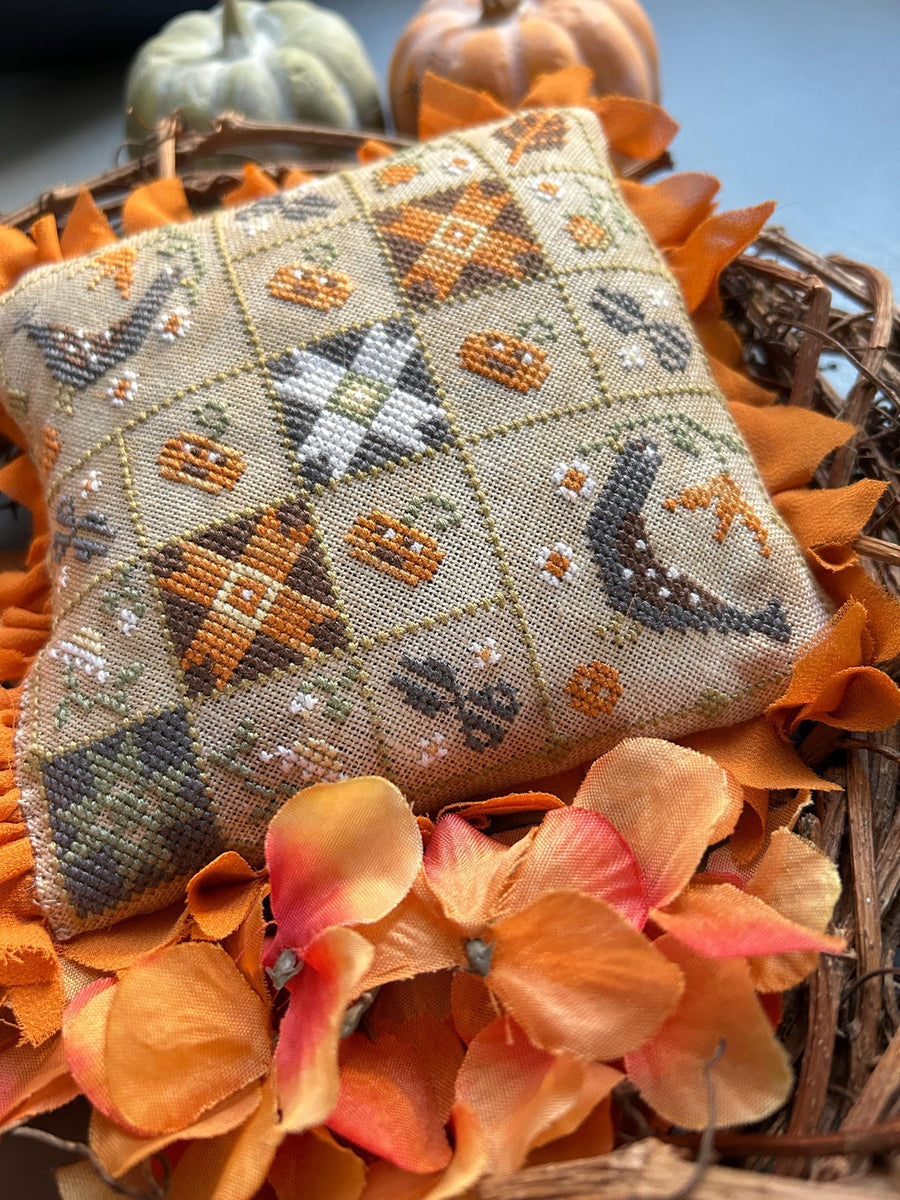 A square decorative pillow with a cross-stitch style autumn pattern, displayed on a wreath surrounded by orange leaves and small pumpkins.