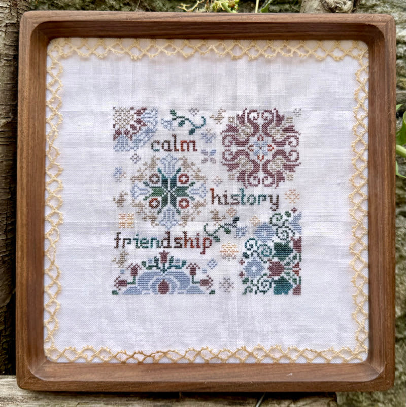 A square framed cross-stitch piece with embroidered text reading 'calm history friendship' surrounded by floral and geometric motifs, set in a wooden frame against an outdoor background.
