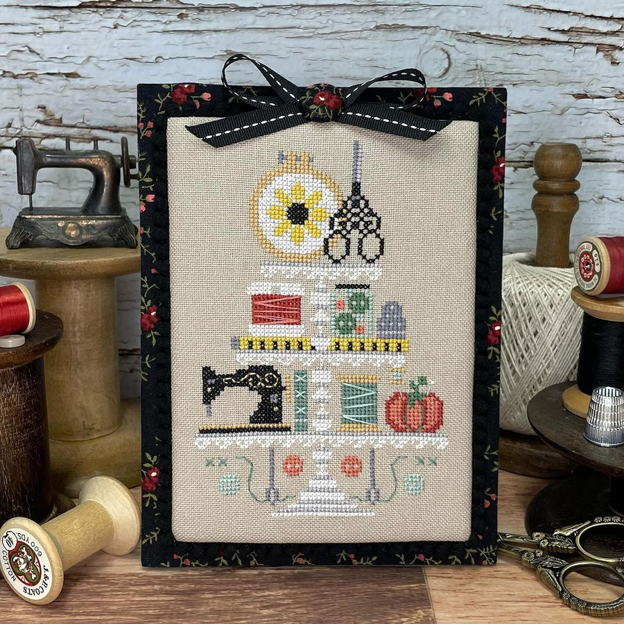 Framed cross-stitched art with sewing-themed design on a rustic wooden background.