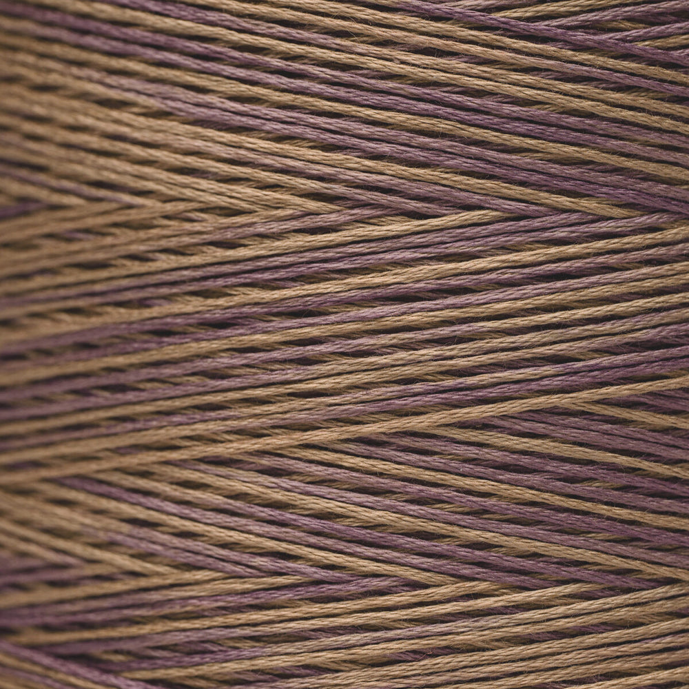 Close-up of variegated yarn wound in diagonal rows, showing alternating strands of warm tan/beige and muted plum-purple.