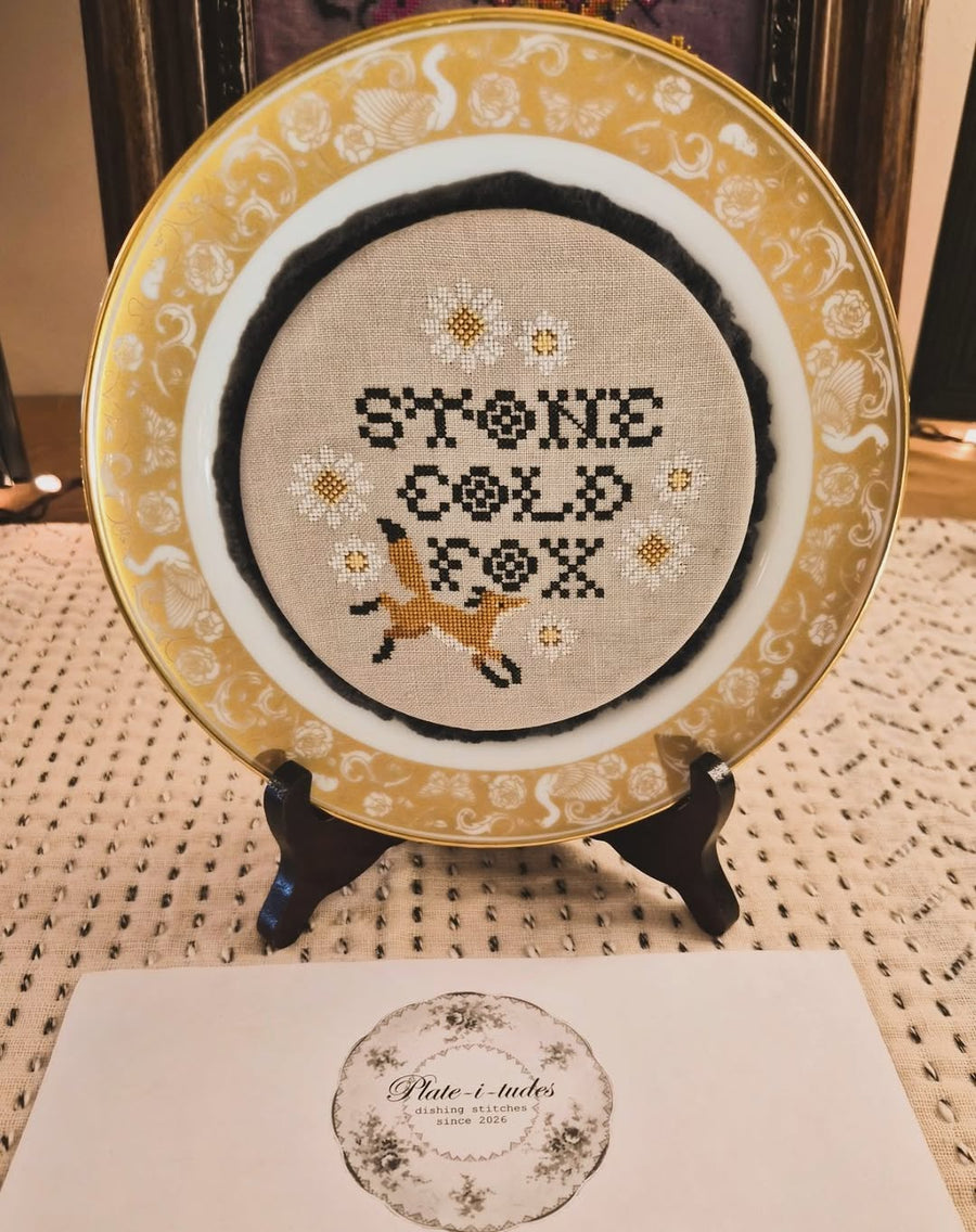 Decorative plate with cross-stitched design of a fox and text on a stand.