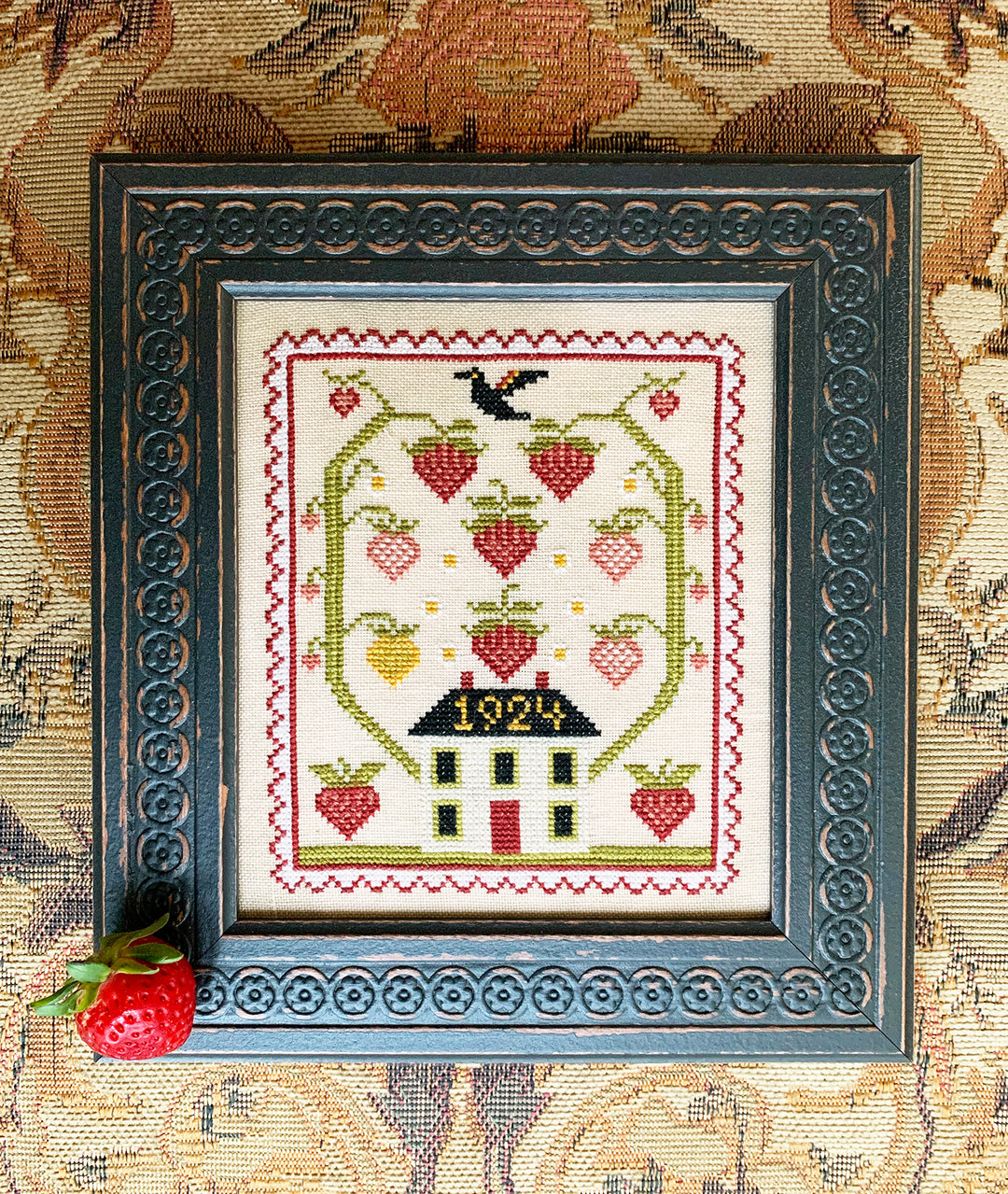 Framed cross-stitched piece with a house and strawberries on a patterned background