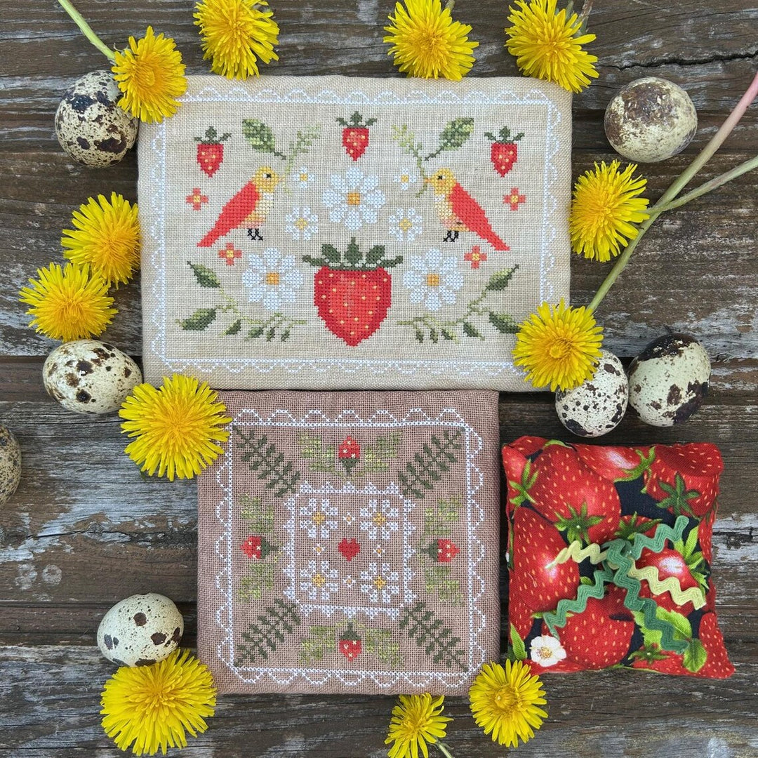 Cross-stitched patterns with strawberries, birds, and flowers on fabric, surrounded by yellow dandelions and speckled eggs on a wooden surface.