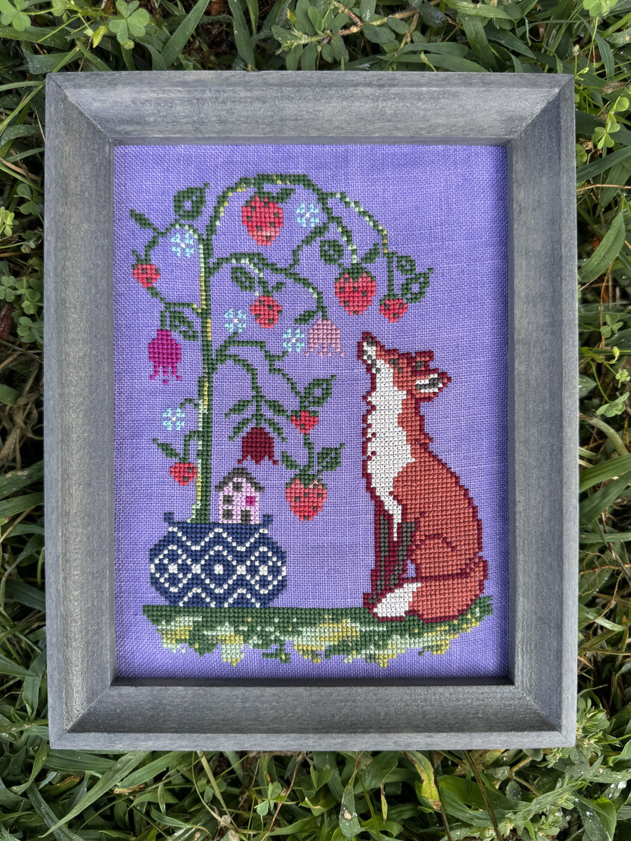A framed cross-stitch artwork featuring a fox and a berry tree, placed on green grass. The artwork has a lavender background with a red fox, green tree, red berries, and a blue patterned pot.