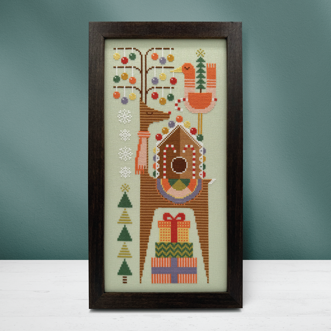 A framed vertical needlepoint or cross-stitch design featuring a stylized tree with colorful ornaments, a bird, a birdhouse, snowflakes, Christmas trees, and wrapped gifts, displayed against a muted blue-green wall on a white surface.