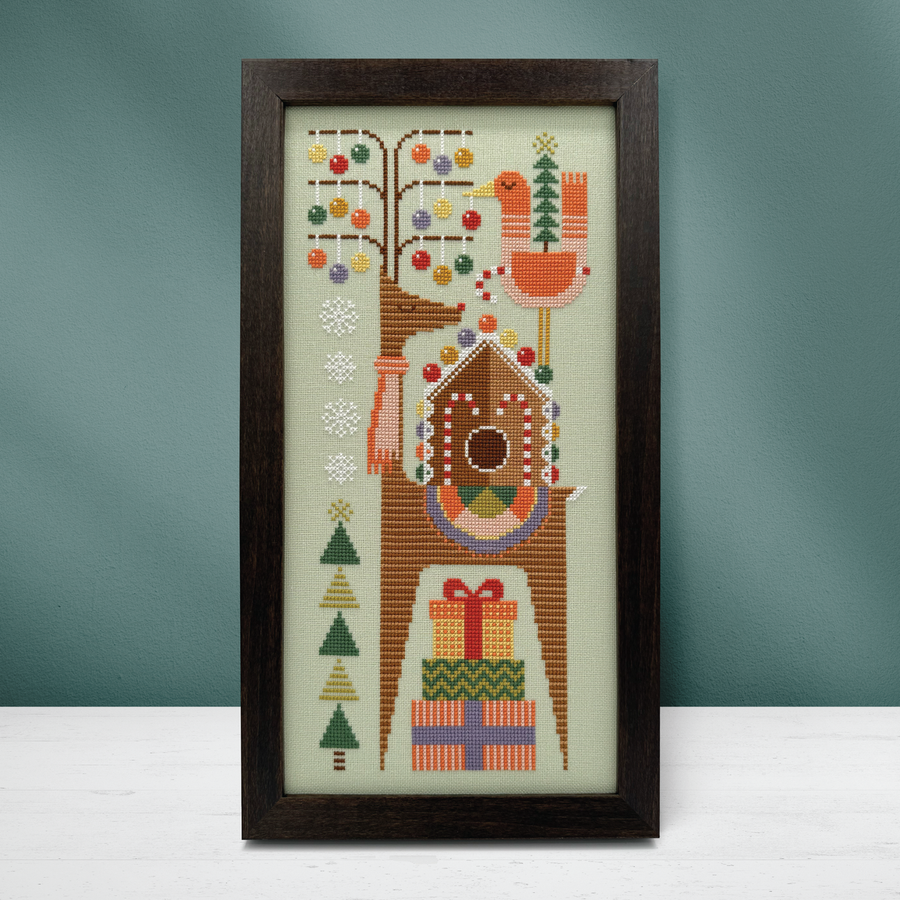 A framed vertical needlepoint or cross-stitch design featuring a stylized tree with colorful ornaments, a bird, a birdhouse, snowflakes, Christmas trees, and wrapped gifts, displayed against a muted blue-green wall on a white surface.