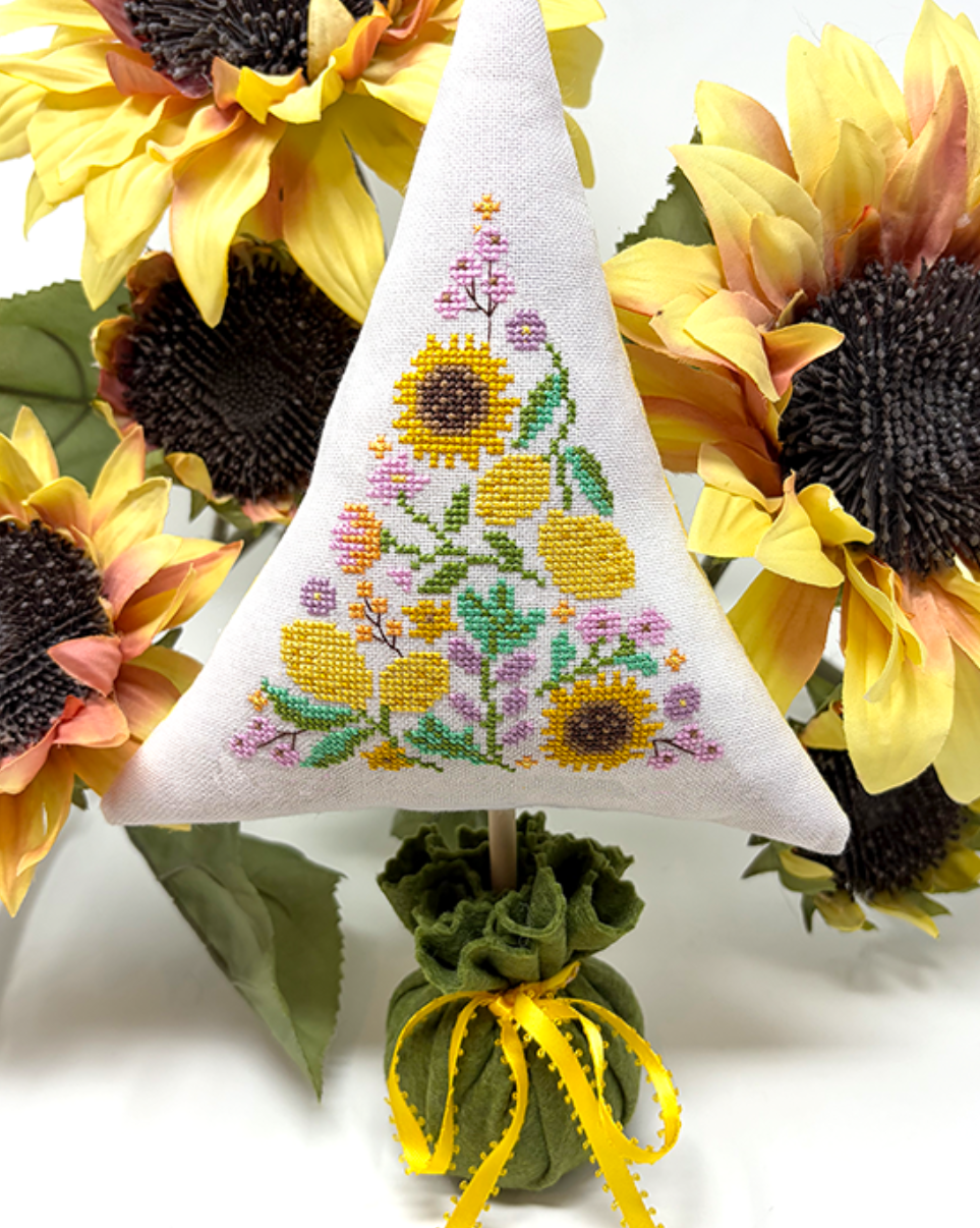 A triangular white embroidered pillow or cushion featuring a sunflower and floral pattern, displayed upright among large yellow sunflowers on a white background.