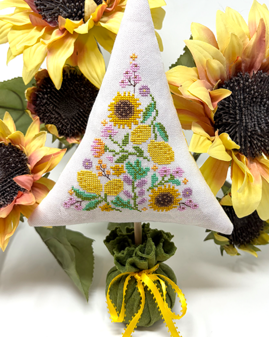A triangular white embroidered pillow or cushion featuring a sunflower and floral pattern, displayed upright among large yellow sunflowers on a white background.