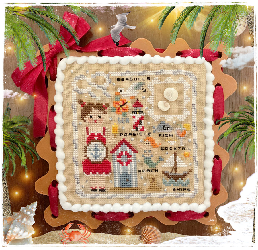 A framed cross-stitch artwork featuring nautical and seaside motifs, including a seagull, lighthouse, fish, cocktail fish, beach, ships, and text labels. The stitched panel is mounted on a scalloped wooden frame with red ribbon accents and surrounded by decorative holiday-style props such as greenery, shells, and warm lights.