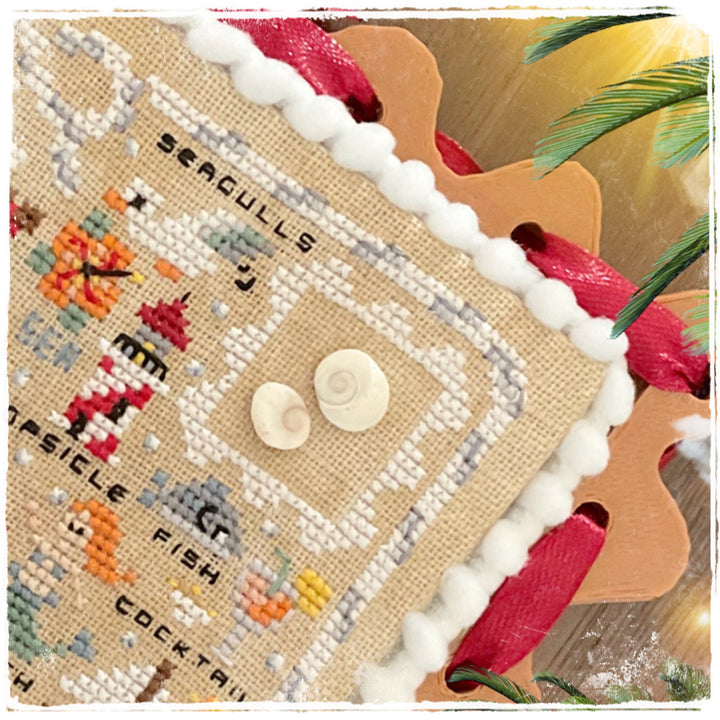 A close-up of a handmade cross-stitch sampler featuring nautical themes, including 'Seagulls', 'Ship'slice', and 'Cocktail Fish'. The sampler is made of natural linen fabric with white pom-pom trim, two small seashell buttons, and colorful embroidered details.