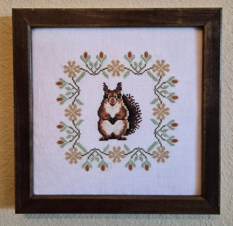 Framed cross-stitched picture of a squirrel with floral patterns on a white background.