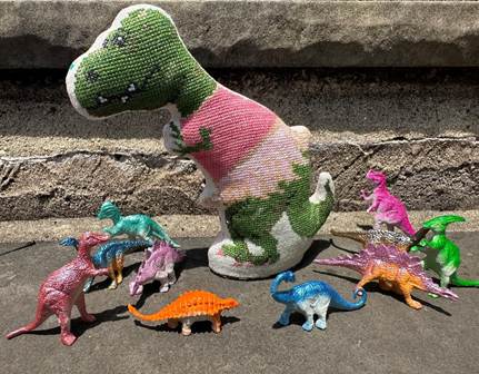 A handmade crocheted T-rex toy with a pink and green patterned body, standing on a concrete surface next to several small colorful dinosaur figurines.