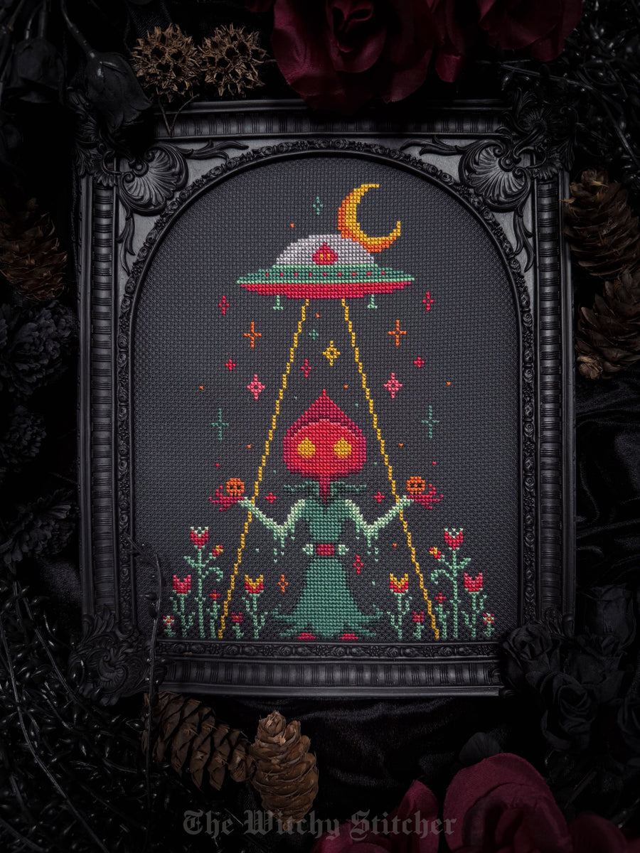 A framed gothic cross-stitch artwork featuring a green cloaked figure with glowing red eyes under a red and green UFO with a yellow crescent moon, surrounded by flowers and stars on a dark gray fabric background. The piece is displayed in an ornate black frame decorated with dark red roses and pinecones, with the text 'The Witchy Stitcher' visible at the bottom.