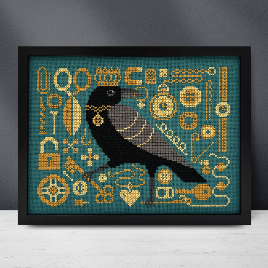A framed cross-stitch artwork featuring a black raven with gold jewelry and motifs on a teal background, displayed against a dark gray wall and light surface.