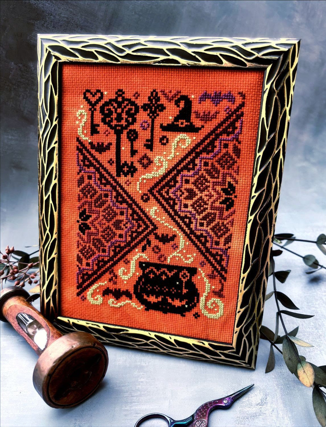 A framed cross-stitch artwork with a Halloween theme, featuring a bright orange fabric background, black and red embroidered motifs including keys, a witch hat, bats, and a cauldron, set in a decorative gold and black frame. The frame is styled on a gray surface with a wooden spool, embroidery scissors, and greenery.